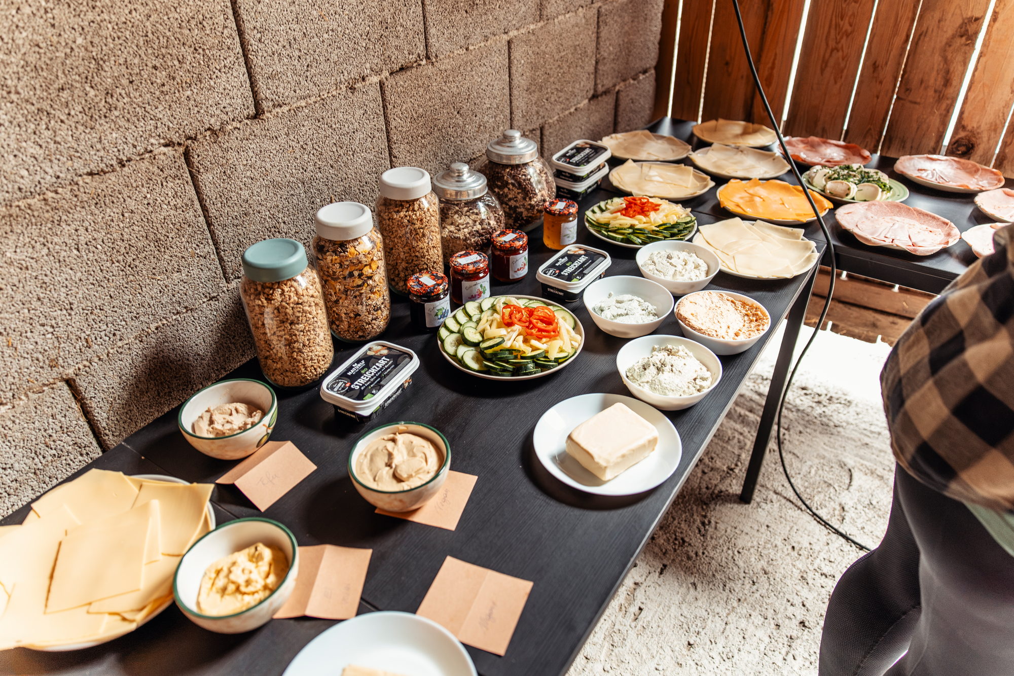 Langer Tisch mit Schalen, Gläsern und Aufstrichen an einer Steinwand, davor Brot und Käse.